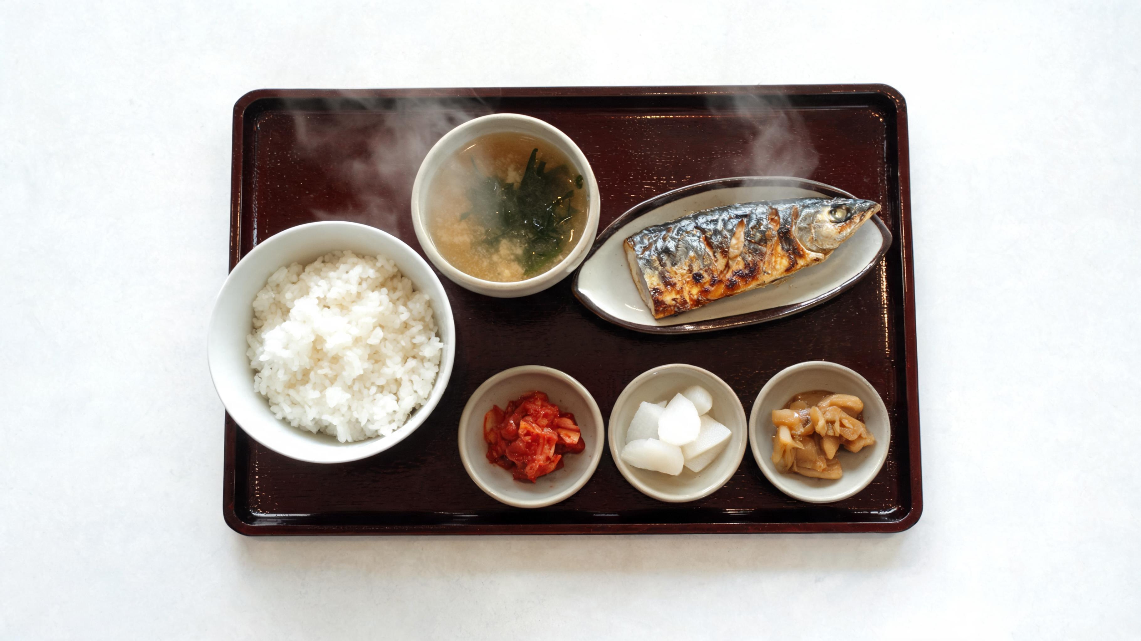 Vista superior de uma bandeja de teishoku laqueada marrom contendo uma tigela de arroz de cerâmica, uma tigela de sopa de missô com wakame flutuando, um pequeno prato de cavala grelhada e três pequenos pratos de picles e vegetais cozidos, vapor subindo suavemente, em uma mesa branca simples.